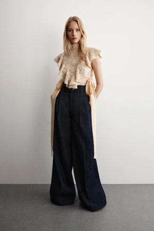 Blonde model wears a tan ruffled blouse and wide, high-waisted, dark-blue pants against a white background in a fashion studio setting.