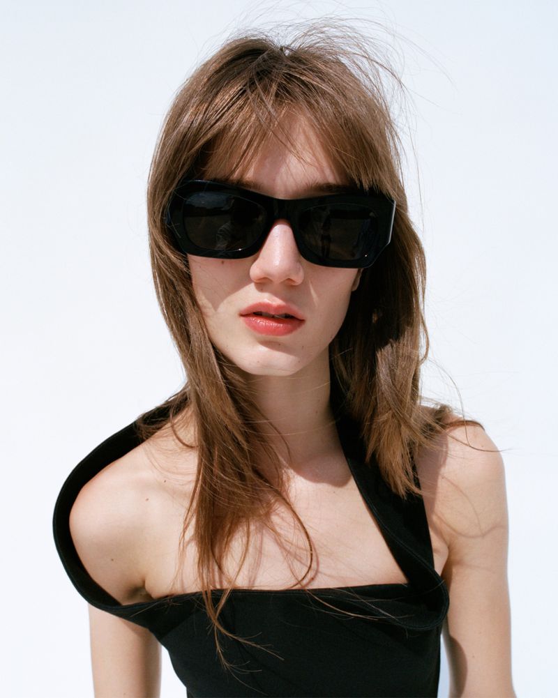 Portrait of a brunette woman with long hair wearing sunglasses and a black top against a white background in a studio setting.