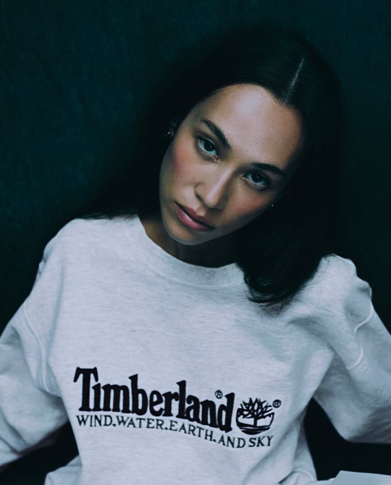 Portrait of a woman wearing a gray Timberland sweatshirt, looking at the camera against a dark background. Fashion lifestyle editorial.
