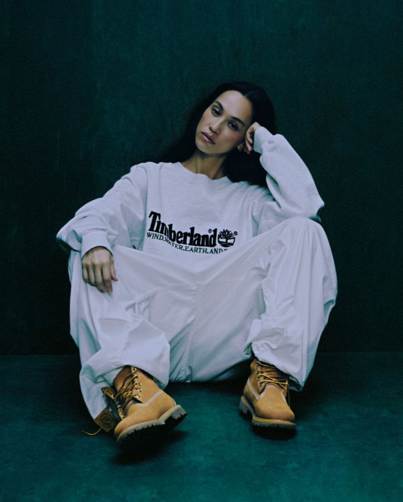 An adult woman sits on the floor wearing a white long-sleeved shirt with a Timberland logo and white pants, along with tan work boots.