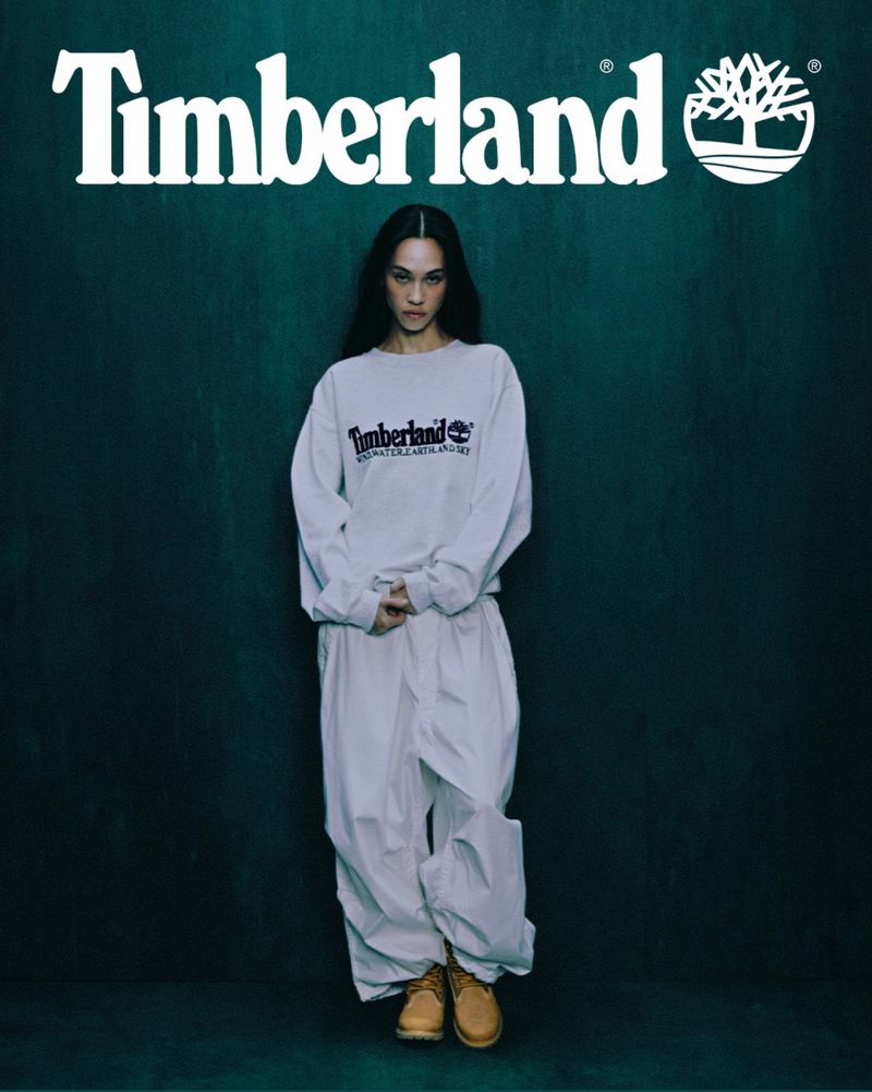 Fashion shot: a slender model in a loose-fitting white outfit and work boots poses against a muted teal background with Timberland logo.