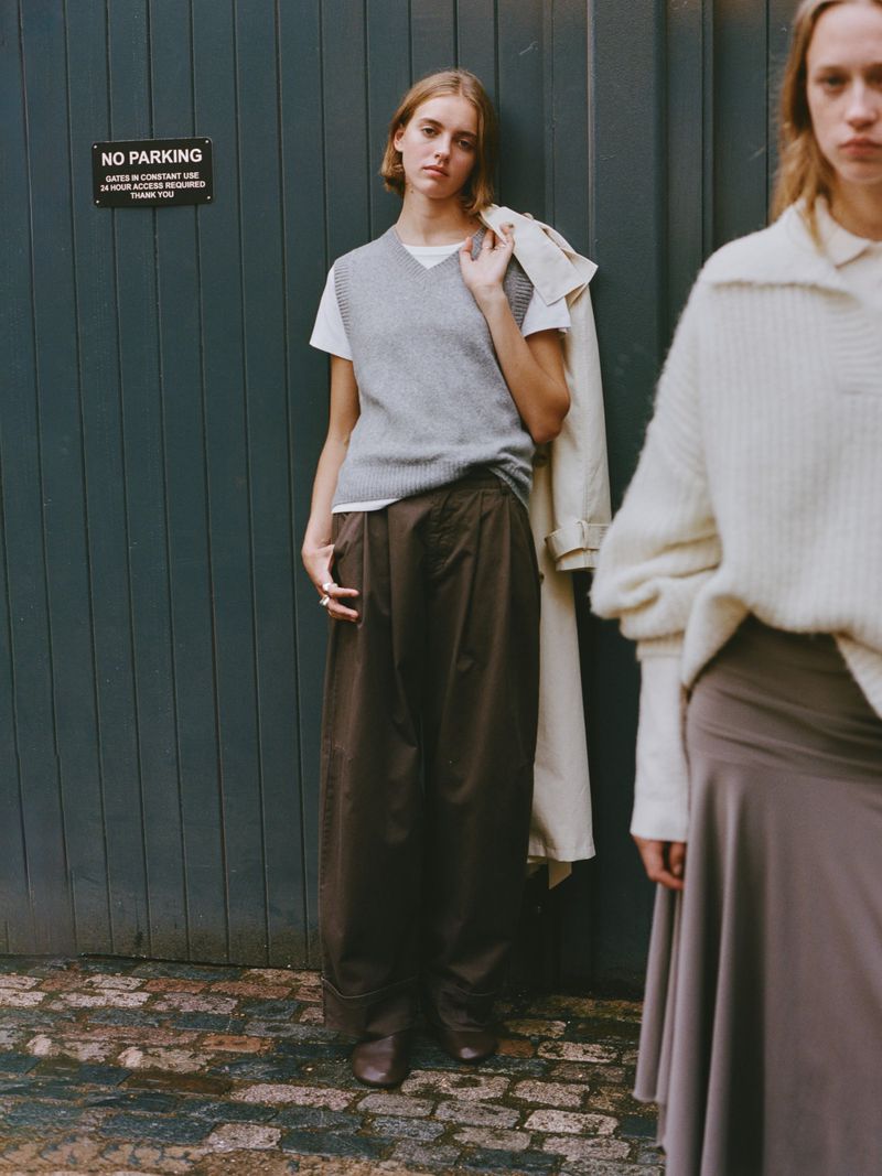 Two women pose against a blue door; one in a grey vest, brown pants and the other in a sweater and skirt, in an outdoor setting.