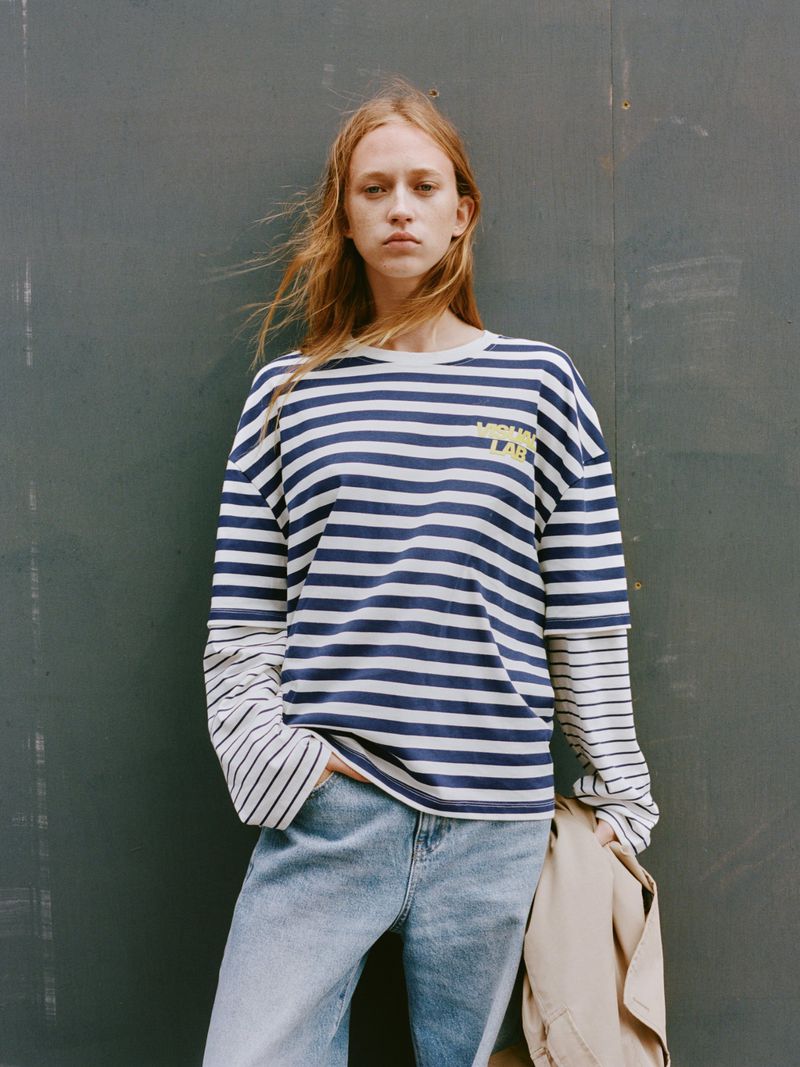 An adult woman poses in a blue-and-white striped long-sleeve shirt and jeans, standing indoors against a gray wall in a medium shot.