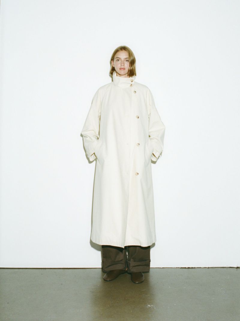 A woman in a light-colored coat and dark trousers stands against a white wall. The coat is buttoned, and her hands are in her pockets.