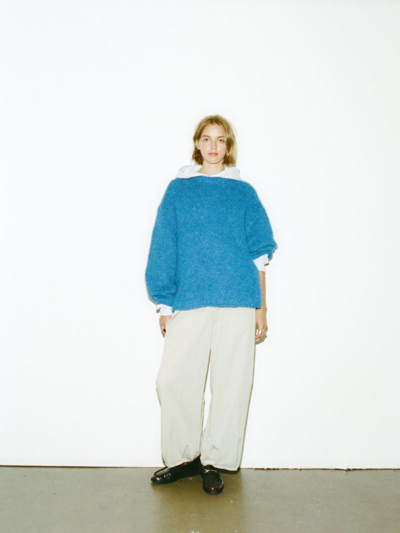 Full shot of woman in a blue knit sweater and baggy pants, standing in front of a white wall with black shoes.