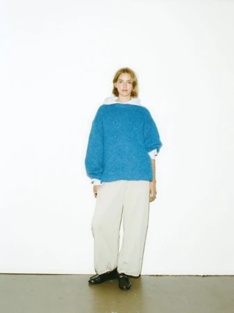 Full shot of woman in a blue knit sweater and baggy pants, standing in front of a white wall with black shoes.