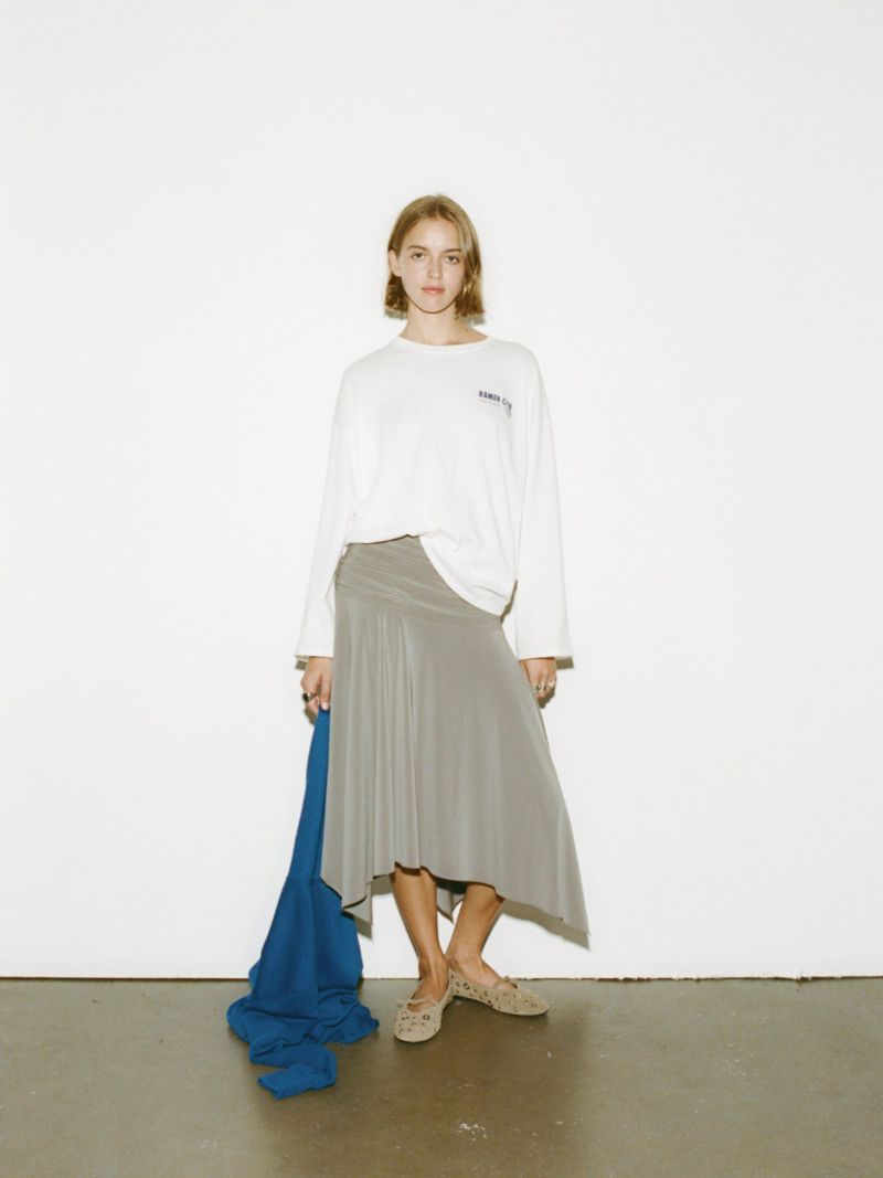 A slender young woman in a long-sleeve white t-shirt and grey skirt poses against a white wall in a fashion or editorial context.