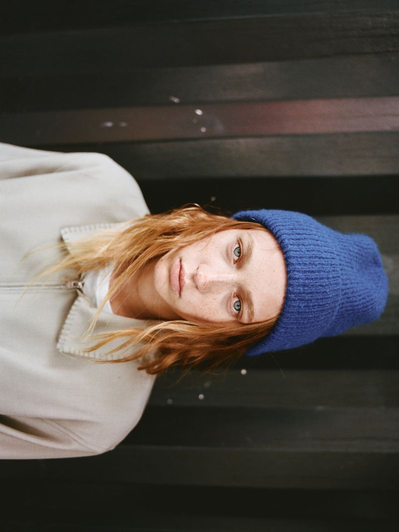 Close-up portrait of a fair-skinned young adult with reddish-brown hair, wearing a blue beanie and light jacket against a dark background.