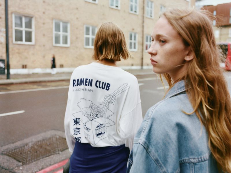 Two young women walk on an urban street. One wears a Ramen Club shirt, the other a denim jacket. A building is in the background.