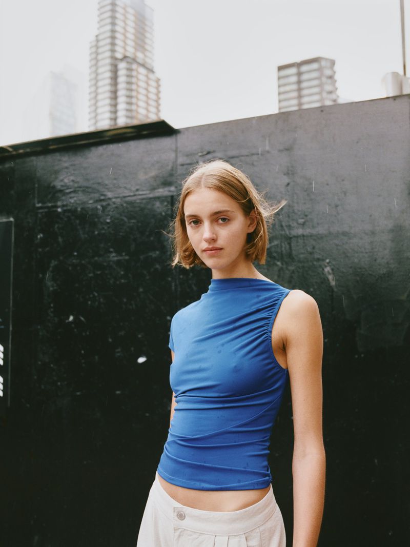 Fashion shot of a blonde woman wearing a blue shirt and light pants stands against a black wall with buildings visible behind.