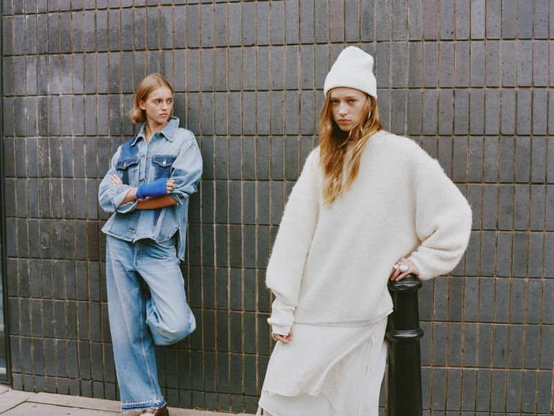 Two blonde-haired women wearing denim and knitwear stand against a gray tiled wall in an outdoor fashion shot.