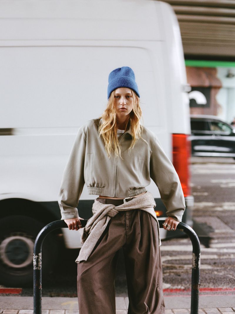 An adult female model poses outdoors in loose, brown pants, a beige jacket, and a blue knit beanie against a white van background.