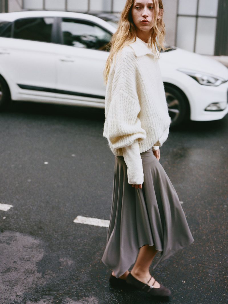 Woman in a cream sweater and gray skirt walks on a wet road with a white car in the background in an outdoor fashion shot.