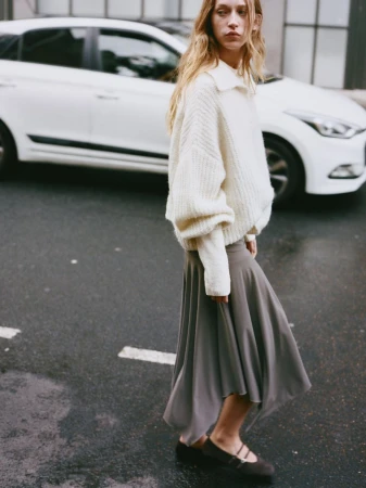 Woman in a cream sweater and gray skirt walks on a wet road with a white car in the background in an outdoor fashion shot.