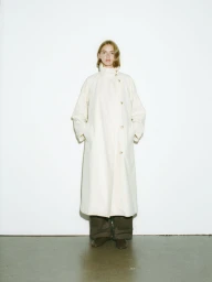 Model standing against a plain white background wearing a long cream coat and dark trousers.