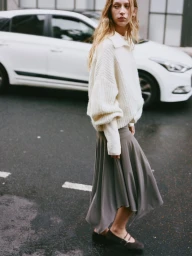 Model in a white knit sweater and grey skirt standing on an asphalt street with a parked white car