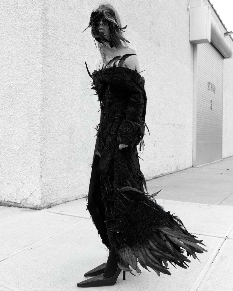 A single adult female fashion model poses in a black feather dress and high heels on a sidewalk in front of a white wall and metal door.