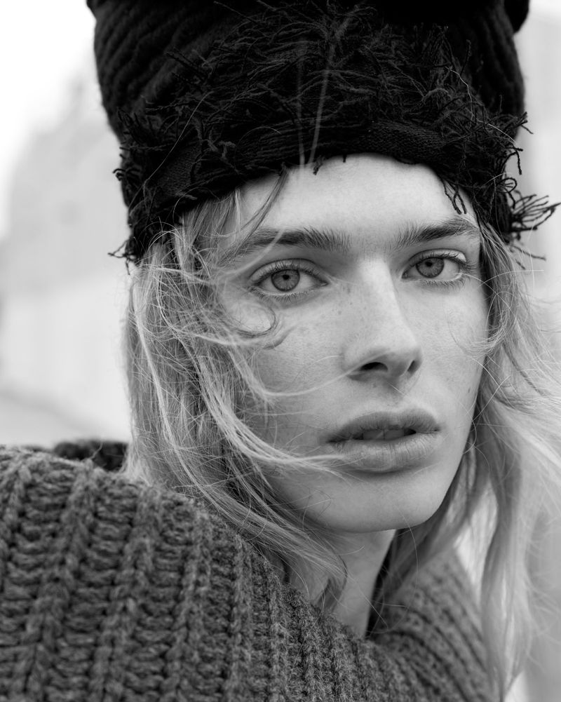 Black-and-white portrait of a blonde adult woman wearing a dark knit hat and sweater against a blurred, neutral-toned backdrop.