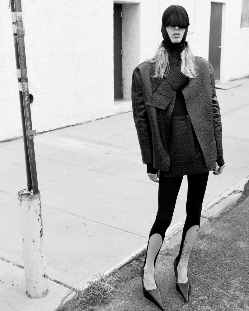 Black-and-white fashion shot of a fair-skinned woman in a black head covering, jacket, skirt, leggings, and heels, standing on a sidewalk.