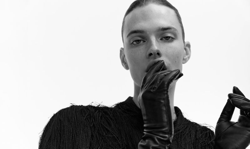 Close-up black and white portrait of a young adult posing with shiny black leather gloves in a minimalist studio fashion shot.