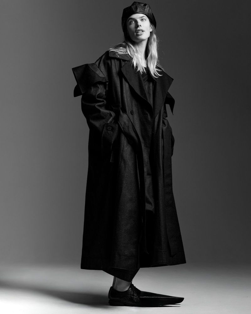 Fashion portrait in black and white featuring a model wearing a long trench coat, beret, and pointed shoes against a grey backdrop.