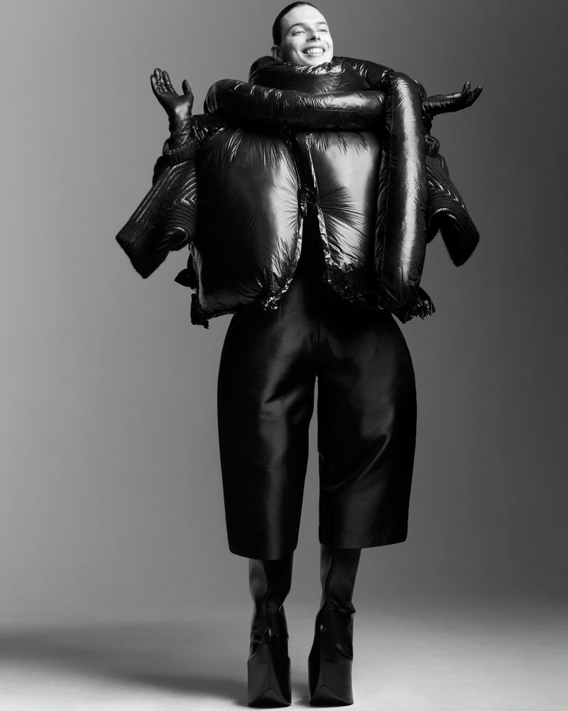 Fashion portrait of woman in black puffer jacket, culottes, gloves, and platform boots against a grey background in studio lighting.