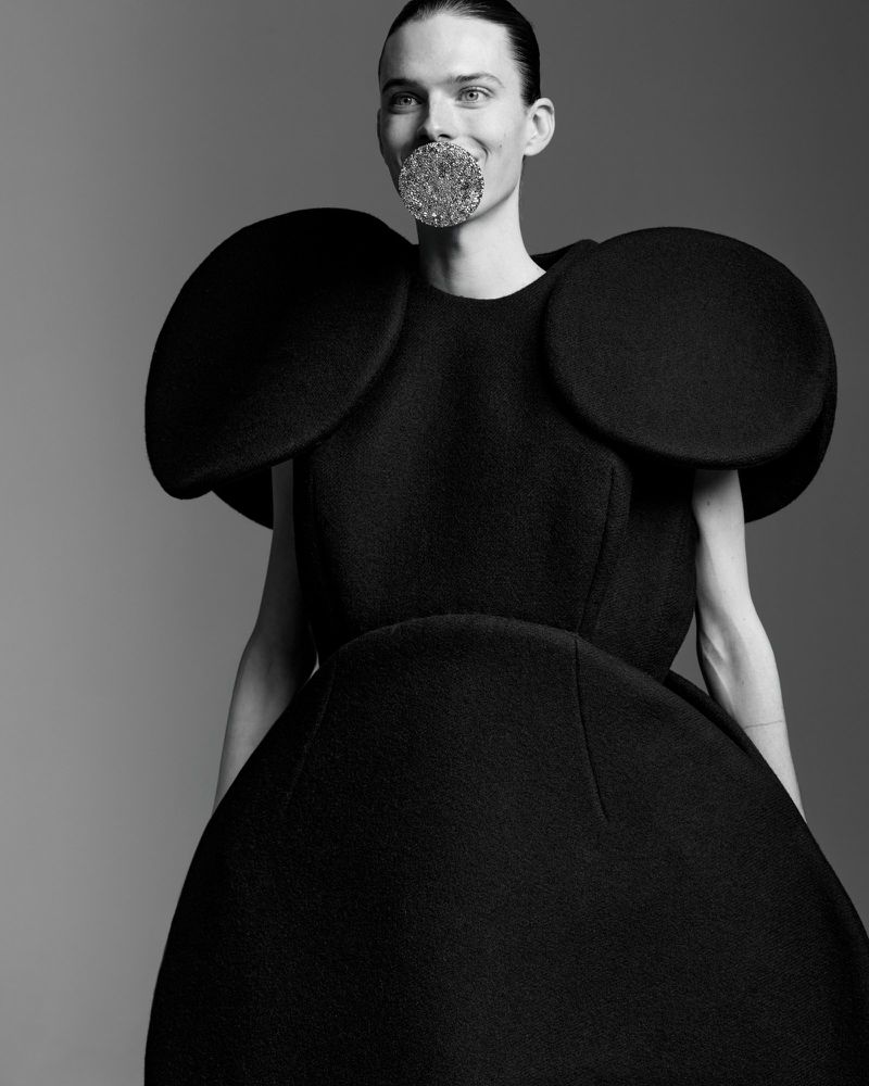 A person in a black sculptural dress with a large round object in their mouth, posed against a gray backdrop in a studio setting.