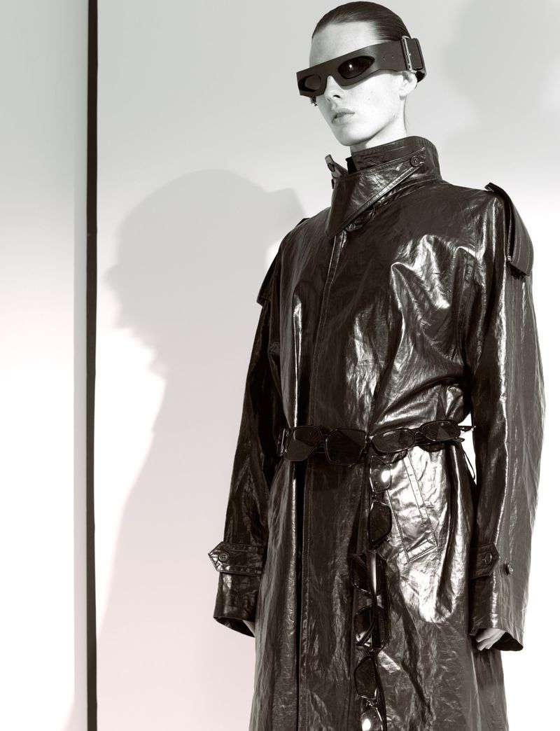 Adult female wearing a long black shiny trench coat and futuristic visor glasses poses in a photo studio against a white background.