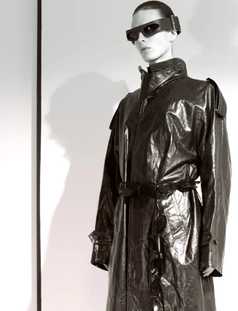 Adult female wearing a long black shiny trench coat and futuristic visor glasses poses in a photo studio against a white background.
