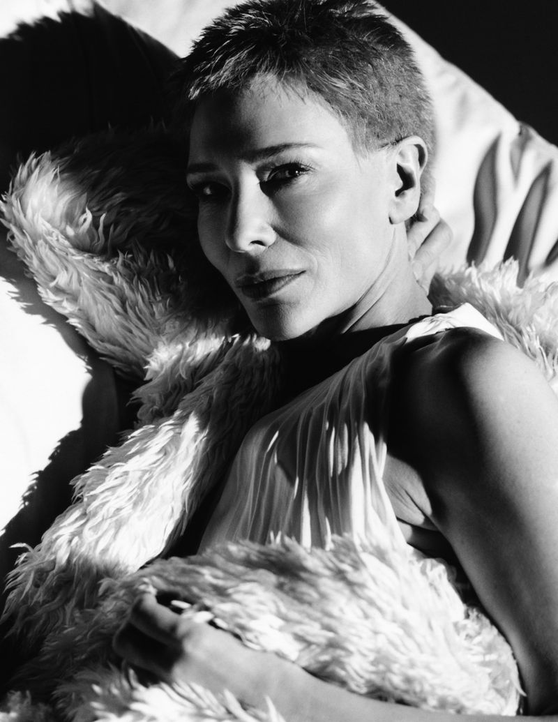 Black-and-white portrait of a smiling adult woman with a pixie cut, lying down, wearing a gown and faux fur shawl indoors.