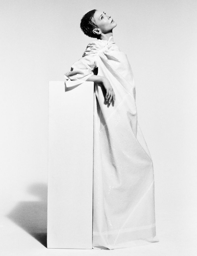 Woman in a white draped gown stands beside a white pedestal in a studio. She looks up, posing in a minimalist setting with soft lighting.