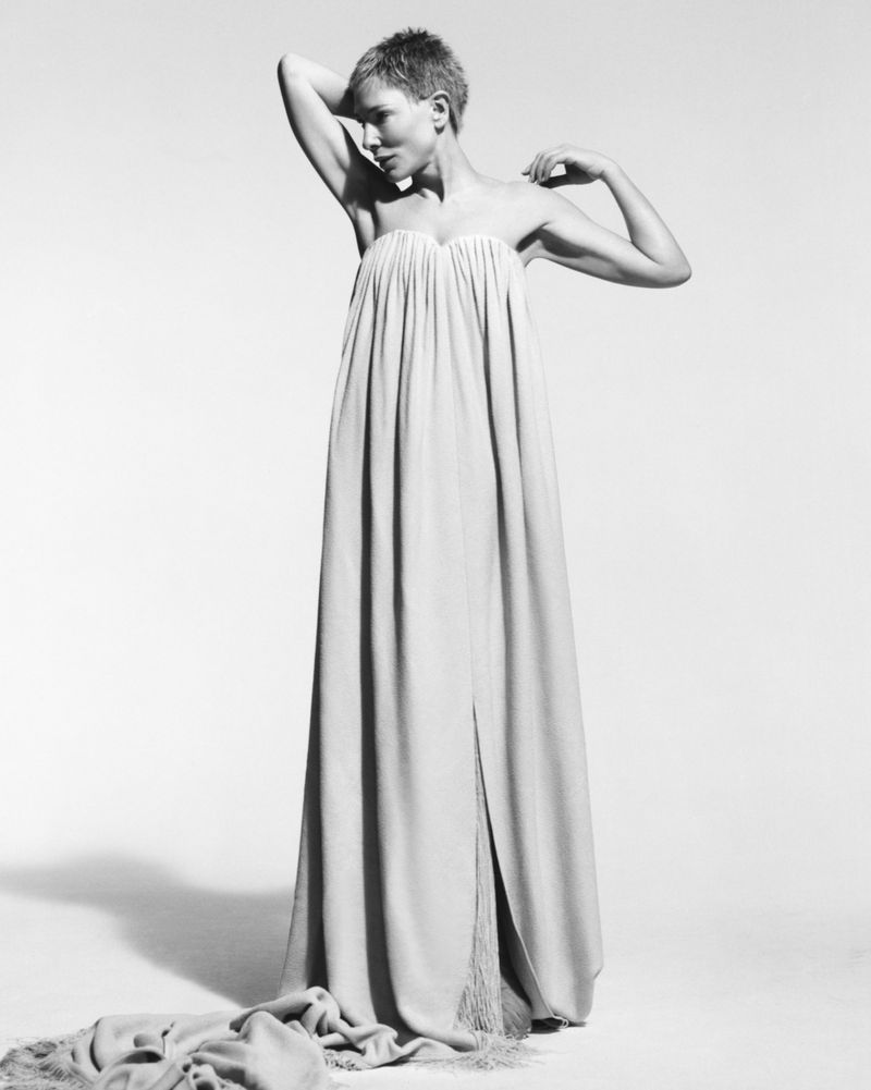 A full-length shot of a solo adult female model with short hair in a long strapless gown, posing in a bright photo studio against a neutral backdrop.