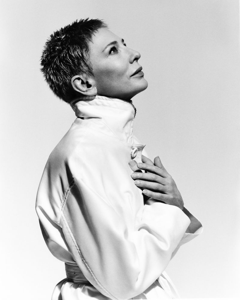 Monochrome portrait of a woman with short hair in a white satin top looking upward, hands clasped at chest, against a plain white backdrop.