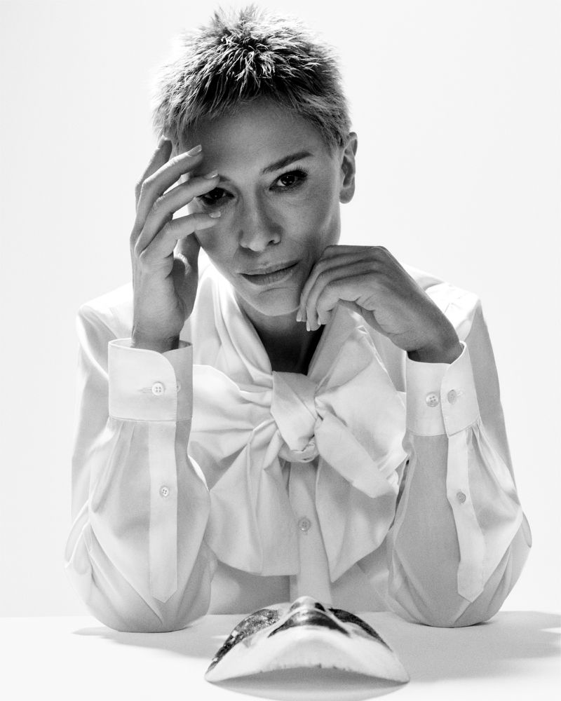 Black and white portrait of an adult female with short hair posing at a table with her hands and a theatre mask.