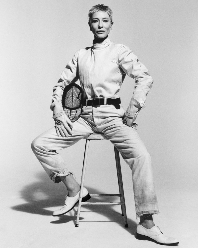 Woman in fencing outfit sits on a stool and holds a fencing mask in a studio setting with neutral background, shot in black and white.