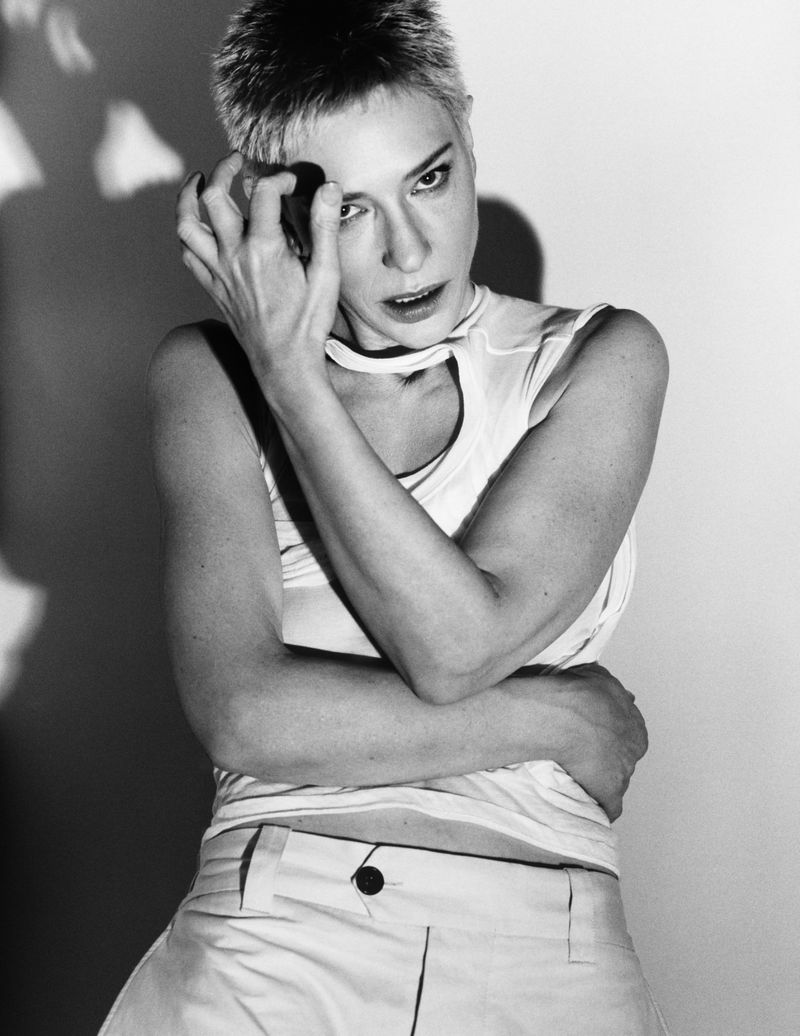 Black and white portrait of a woman with short hair in a white top and pants, posing indoors against a bright wall with a shadow.