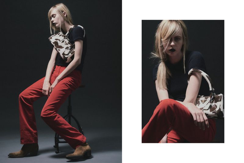 A seated fashion model poses in red pants, a black top, brown boots and holding a cow-print handbag in a studio setting.