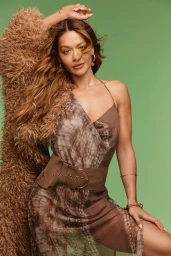 Rita Ora posing against a green backdrop in a brown slip dress with a thick belt and a fluffy coat draped over her shoulder.