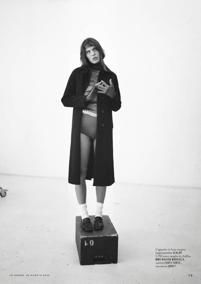 Woman posing in an open black coat, socks, and loafers, standing on a black block in a minimalist white studio with copy text.