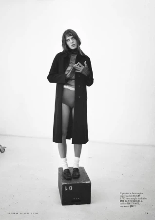Woman posing in an open black coat, socks, and loafers, standing on a black block in a minimalist white studio with copy text.