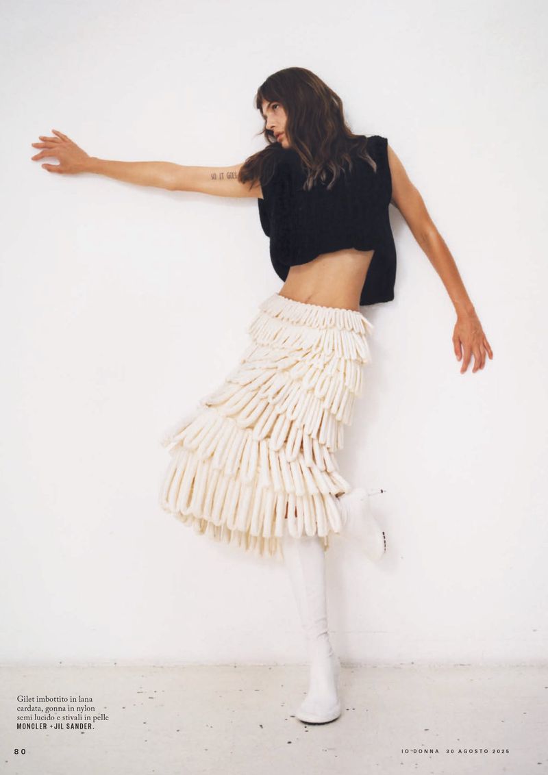 Fashion shoot featuring a slim woman in a black top and textured white skirt, posing against a plain white backdrop in a studio.