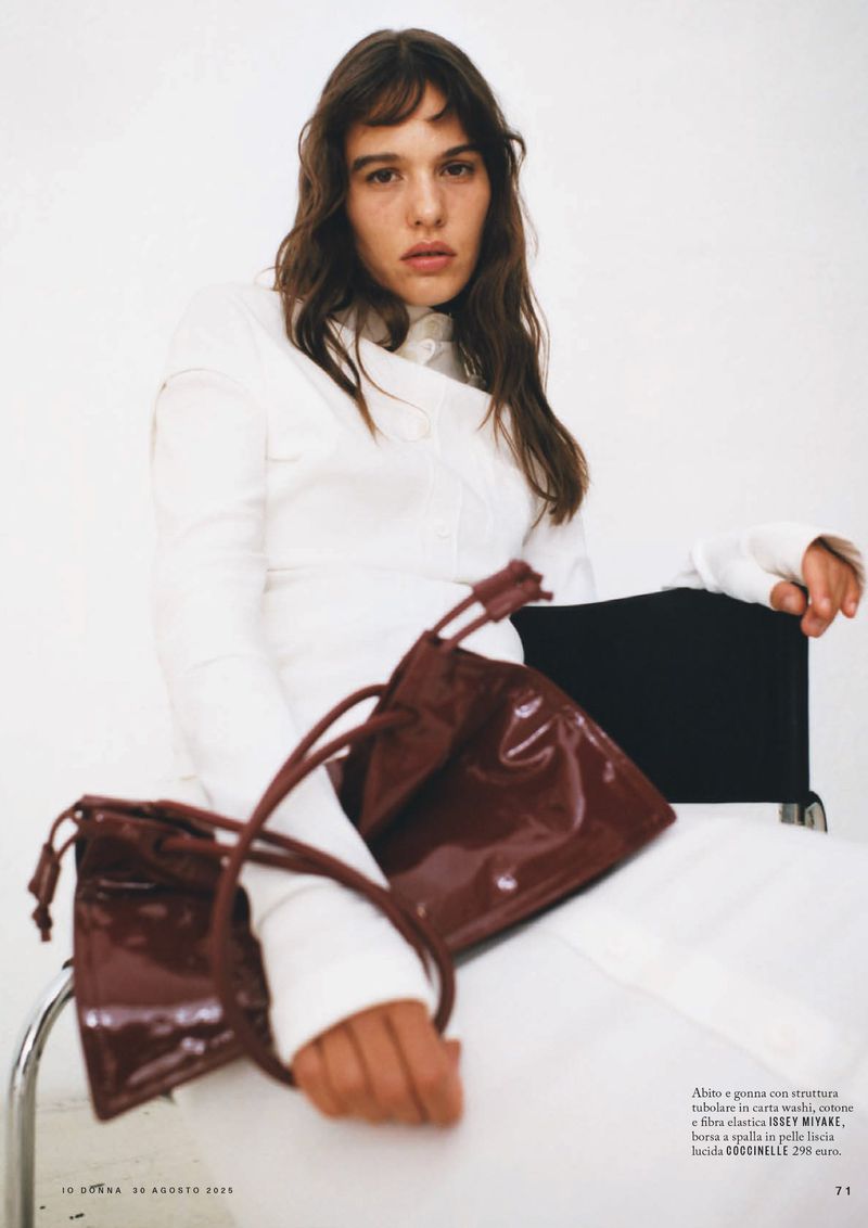 Editorial fashion shot of a brunette woman in a white outfit, seated with two shiny brown purses in an indoor studio setting.