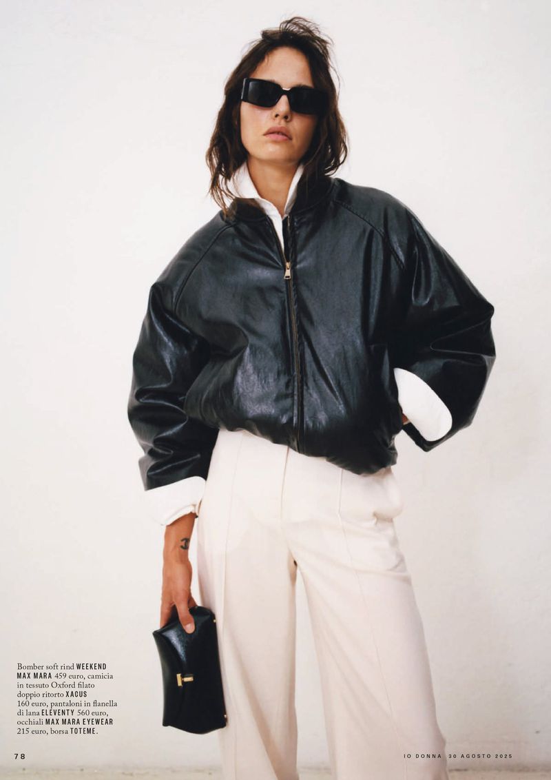 A woman in a black leather jacket, cream pants, and sunglasses stands in front of a white wall, holding a black eyeglasses case.