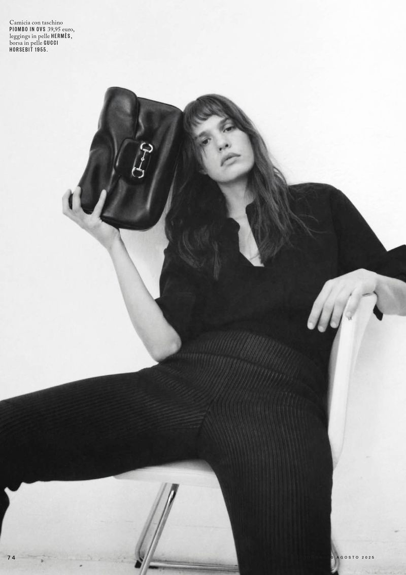Black and white portrait of a woman in black attire seated on a chair, holding a black leather handbag with metal hardware.