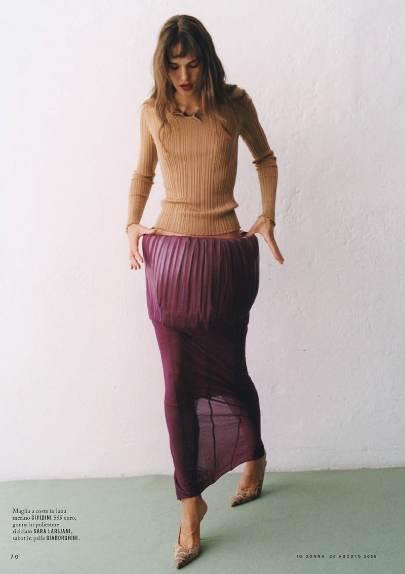 A woman in a light brown ribbed long-sleeve top and a long dark pink pleated skirt poses indoors against a white wall with patterned heels.