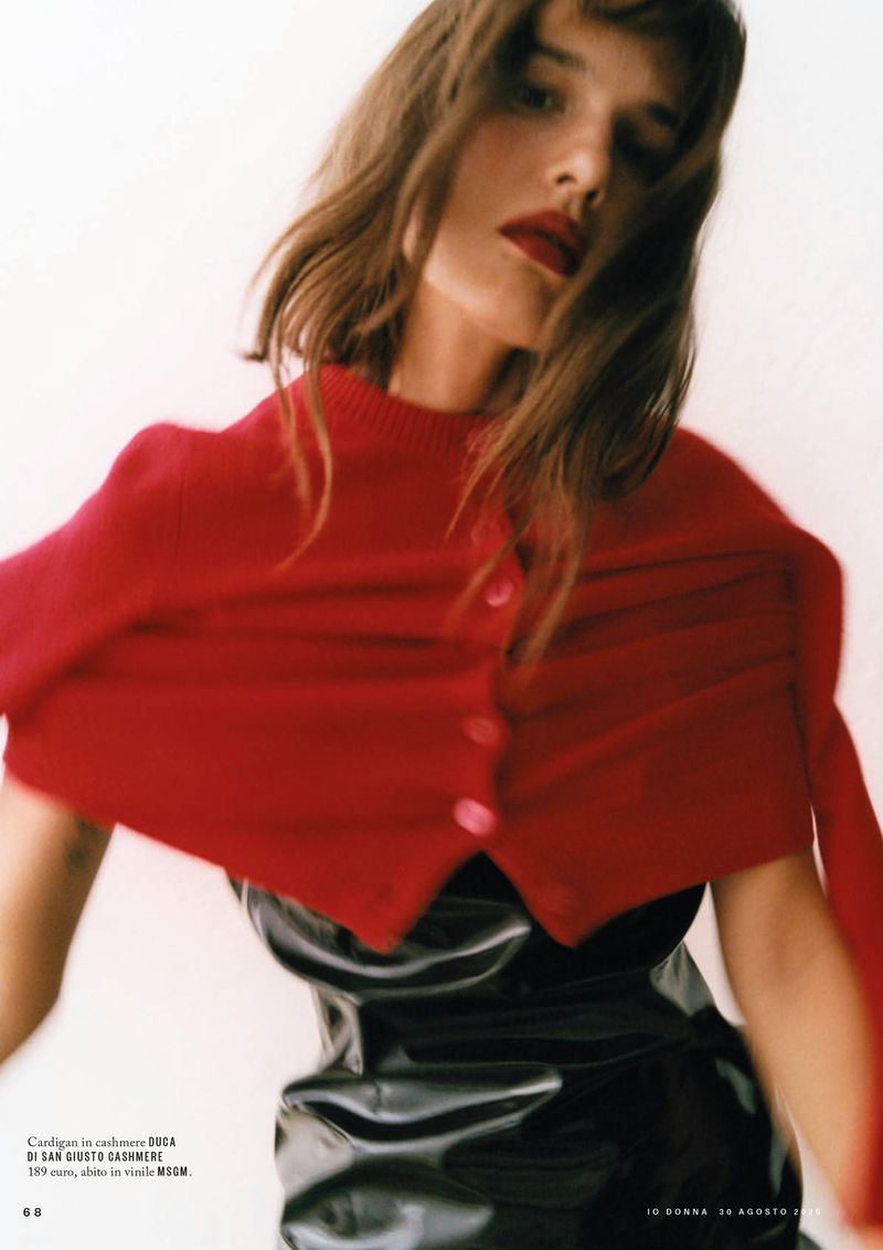 Fashion editorial shot of a brunette woman in a red cardigan and black dress against a white background, with red lipstick.