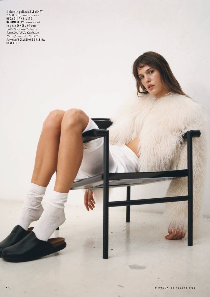 A young woman with bangs sits in a modern chair wearing a textured jacket, white shorts, socks and mules against a white background.