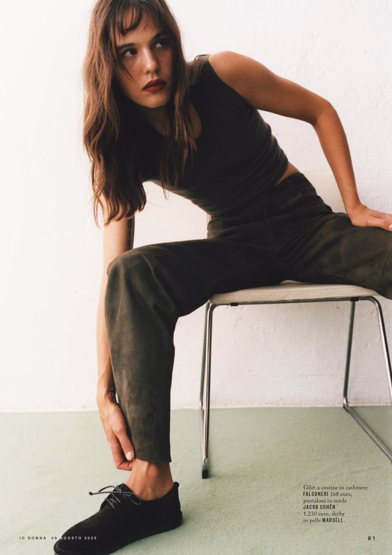 A woman in a dark green tank top and pants sits on a metal chair in front of a white wall with green flooring, showcasing an editorial fashion style.