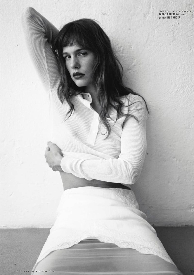 A young woman with dark hair and bangs poses indoors against a wall, wearing a white knit shirt and white lace skirt in a black and white fashion shot.