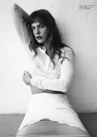 A young woman with dark hair and bangs poses indoors against a wall, wearing a white knit shirt and white lace skirt in a black and white fashion shot.